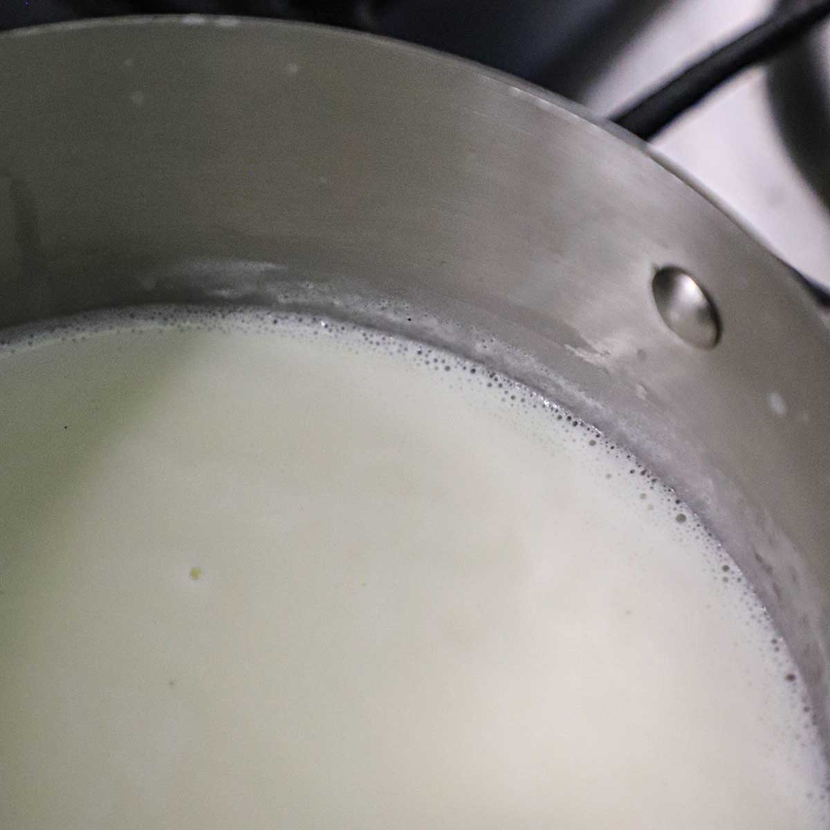 A close-up view of a cream and milk mixture that is just starting to bubble in a saucepan on a gas stove.