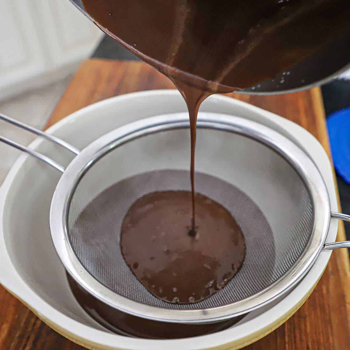A person pouring a chocolate custard through a sieve over a ceramic bowl on a wooden cutting board.