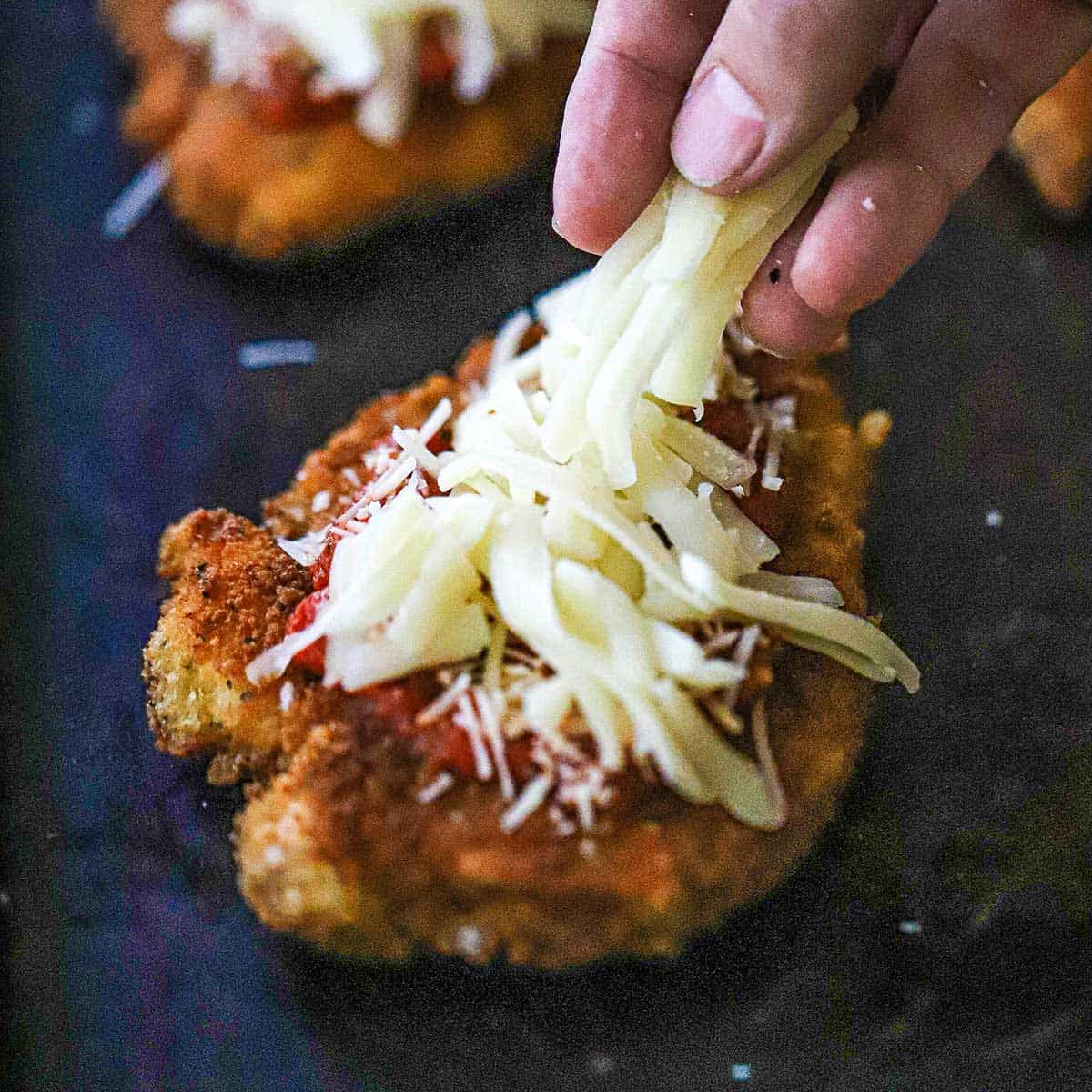 A person placing shredded mozzarella cheese over the top of a lightly breaded and fried chicken cutlet that is also topped with marinara sauce and grated Parmesan cheese.