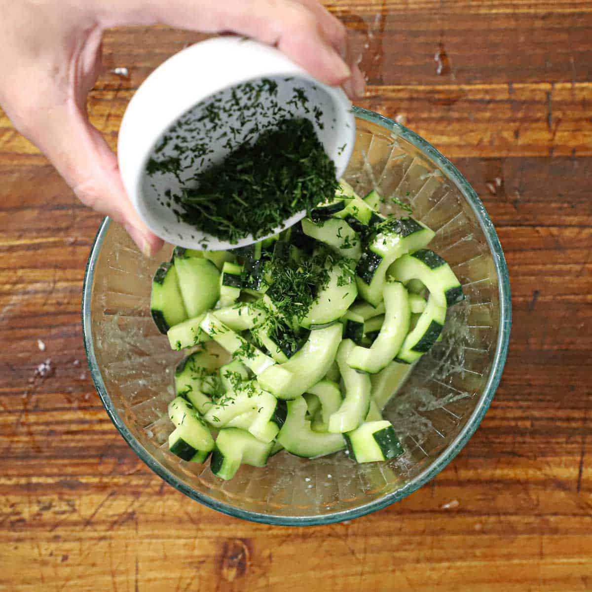 A person transferring chopped fresh dill from a small white bowl into another glass bowl filled with sliced cucumber.