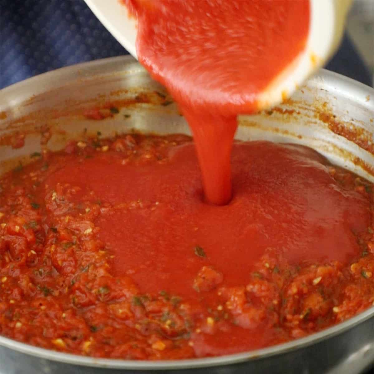 A person pouring tomato sauce from a large ceramic bowl into a large silver skillet filled with simmer crushed tomatoes, herbs, and garlic.