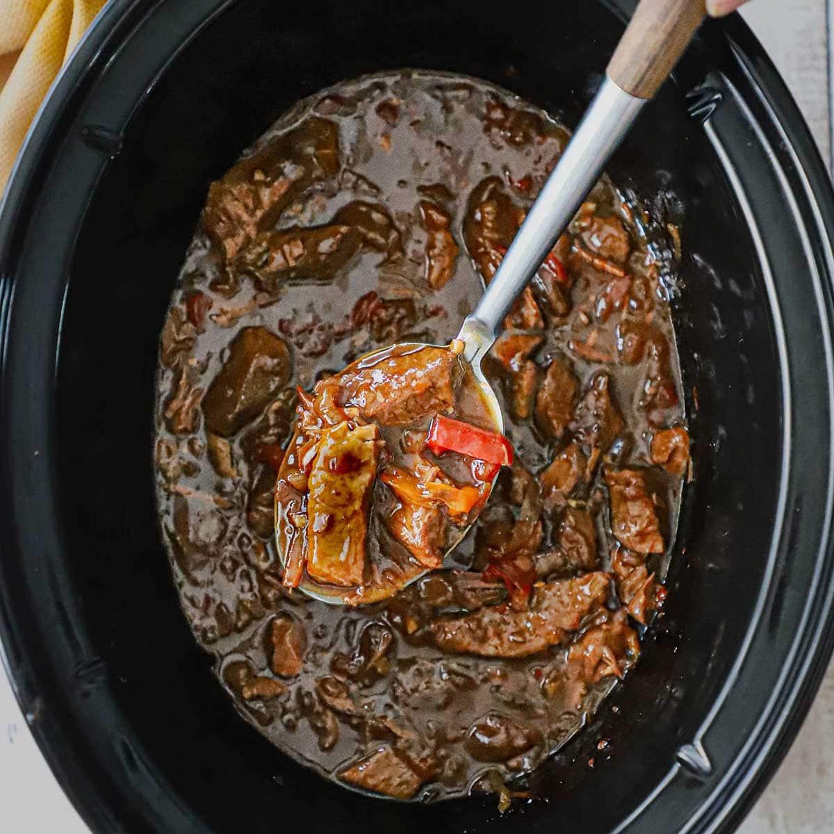 A large serving spoon being used to raise a serving of slow cooker pepper steak from the inside of a large oval slow cooker.