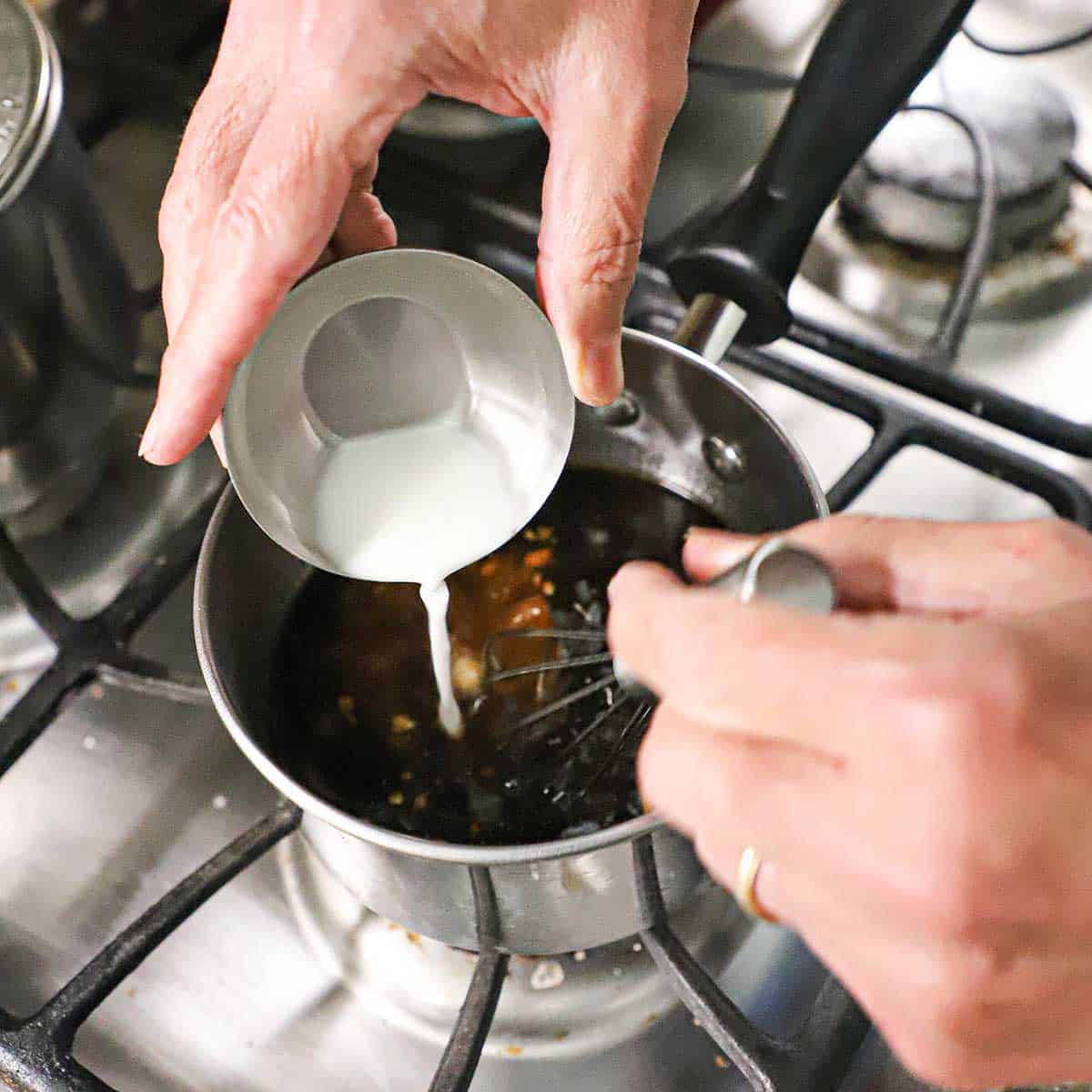 A person pouring a cornstarch slurry from a small silver bowl into a small saucepan filled with a simmering teriyaki sauce on a gas stove.