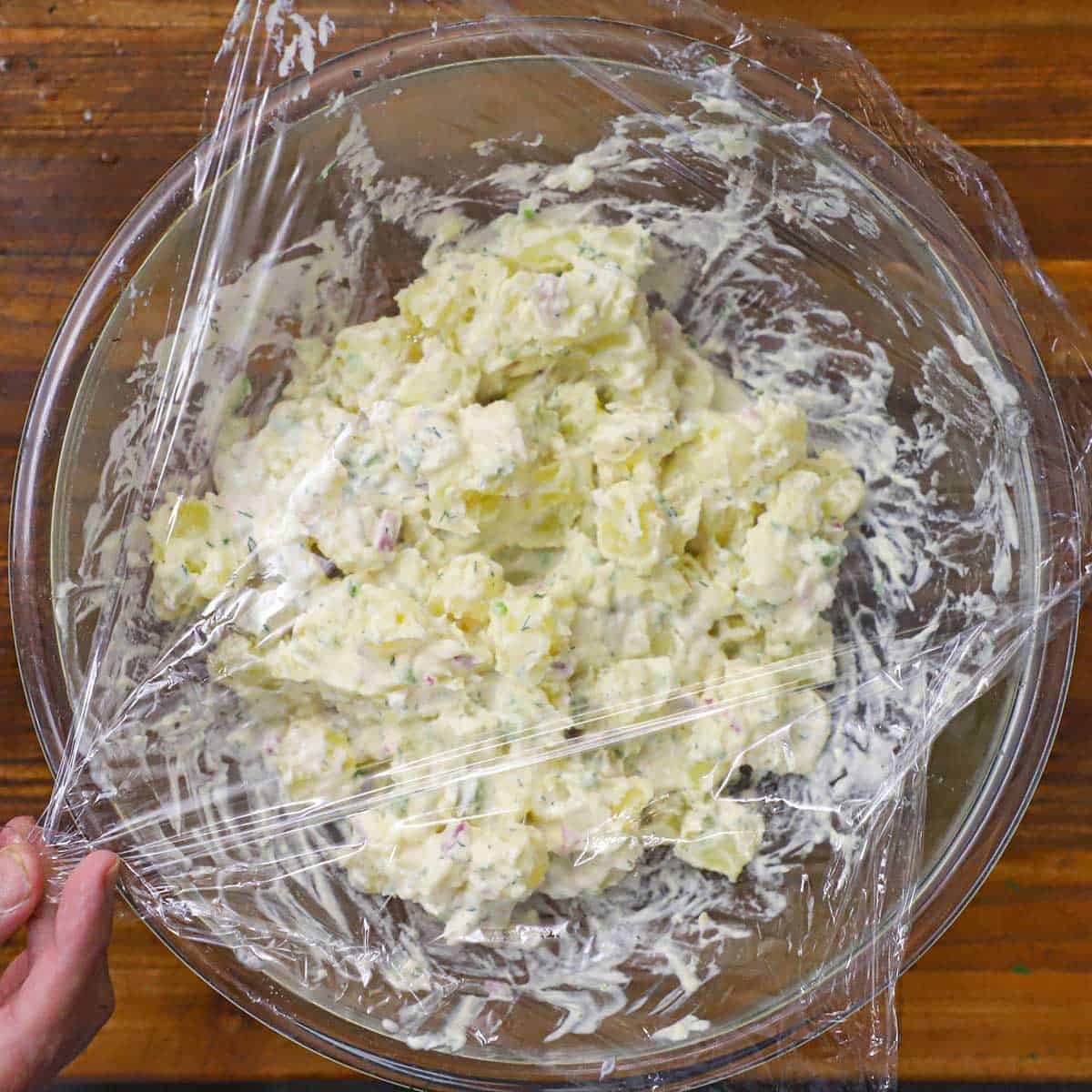 A person spreading plastic wrap over the top of a glass bowl that is filled with freshly prepared best-ever potato salad.