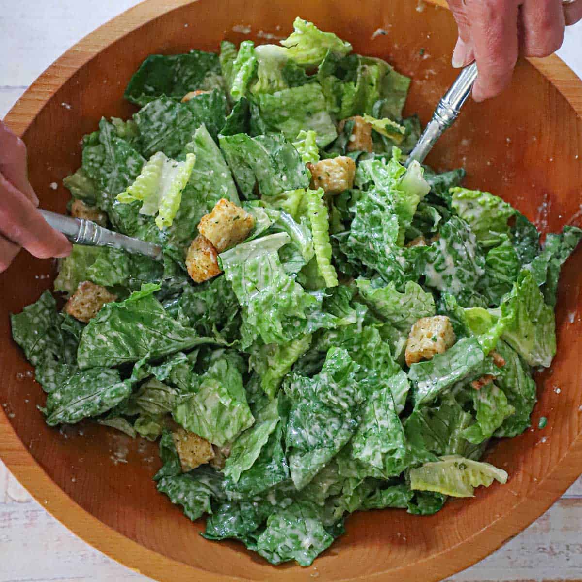 A person using two large silver salad spoons to toss together a classic Caesar salad in a large wooden bowl.