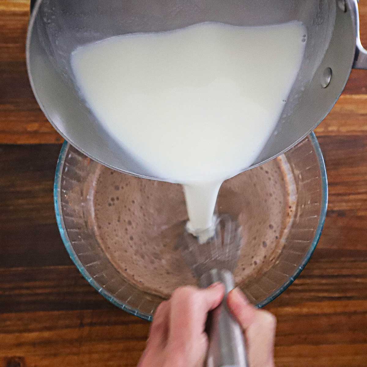 A person pouring a warm cream mixture from a saucepan into another bowl with a chocolate sauce mixture with egg yolks, while whisking to temper the eggs.