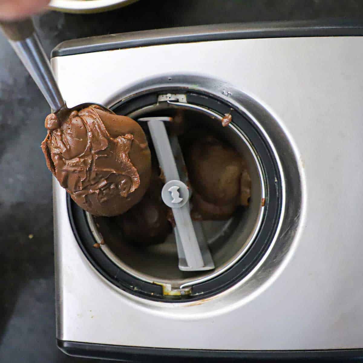 A person using a large spoon to scoop out freshly churned homemade chocolate ice cream that is still soft from the bucket of an electric ice cream.