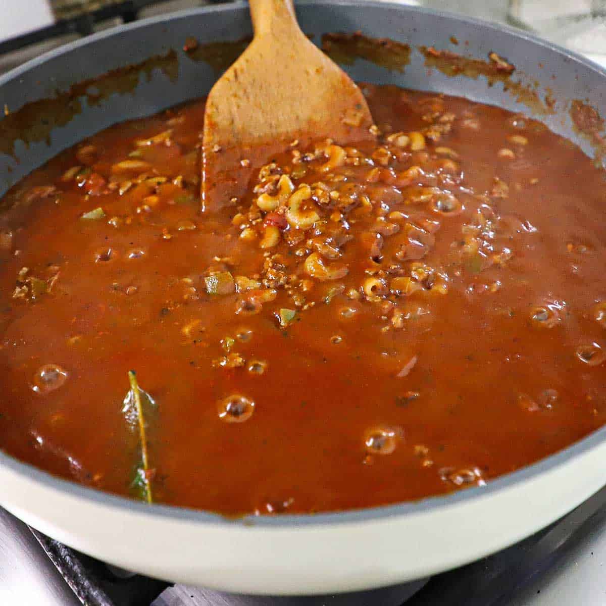 A wooden spatula being used to stir partially cooked elbow macaroni in a large skillet filled with a tomato and beef sauce.