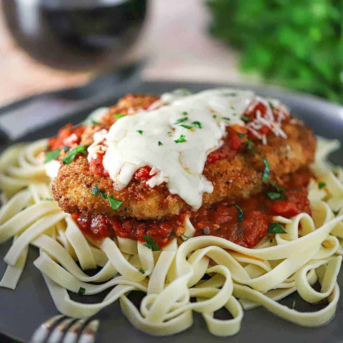 A serving of chicken Parmesan (Parmigiana) resting on a bed of cooked linguine with a small mound of homemade marinara sauce just underneath the cutlet.