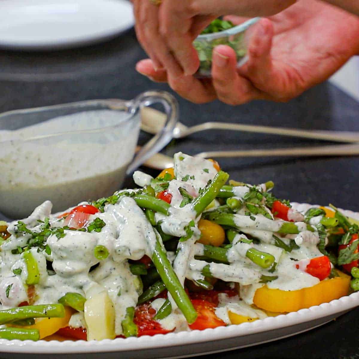 A person using his hand to sprinkle freshly chopped mint over the top of a green bean and tomato salad that has been topped with a scallion buttermilk dressing all on an oval white platter.