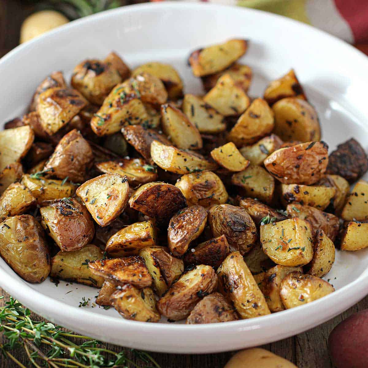 A white serving bowl filled with crispy roasted potatoes with balsamic and herbs with stems of fresh herbs nearby.