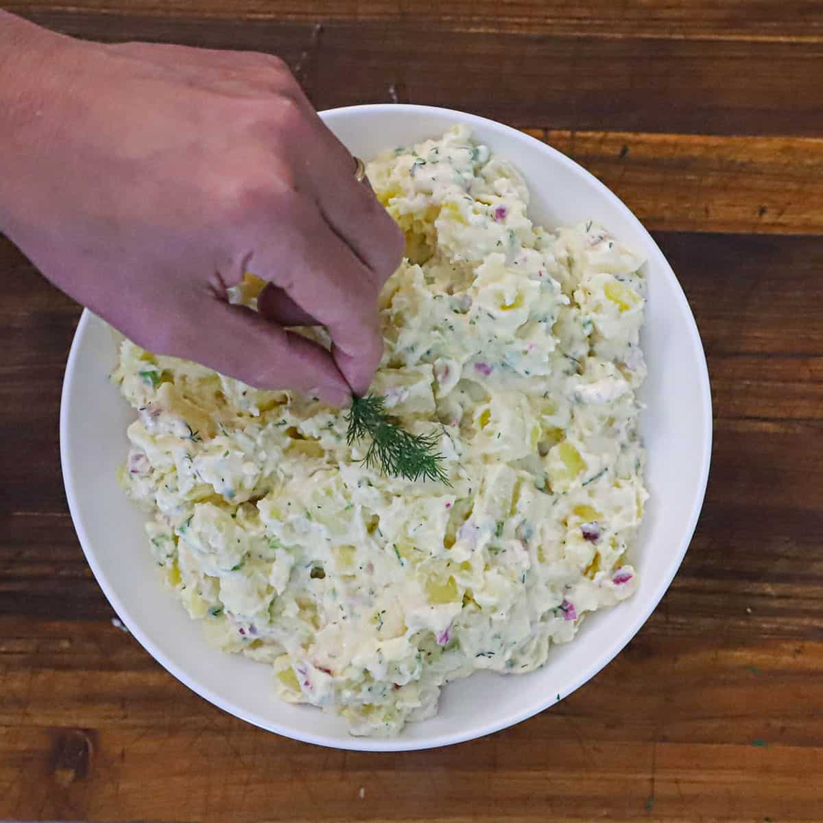 A person placing a sprig of fresh dill on the top of a bowl of best-ever potato salad in a white bowl on a wooden cutting board.