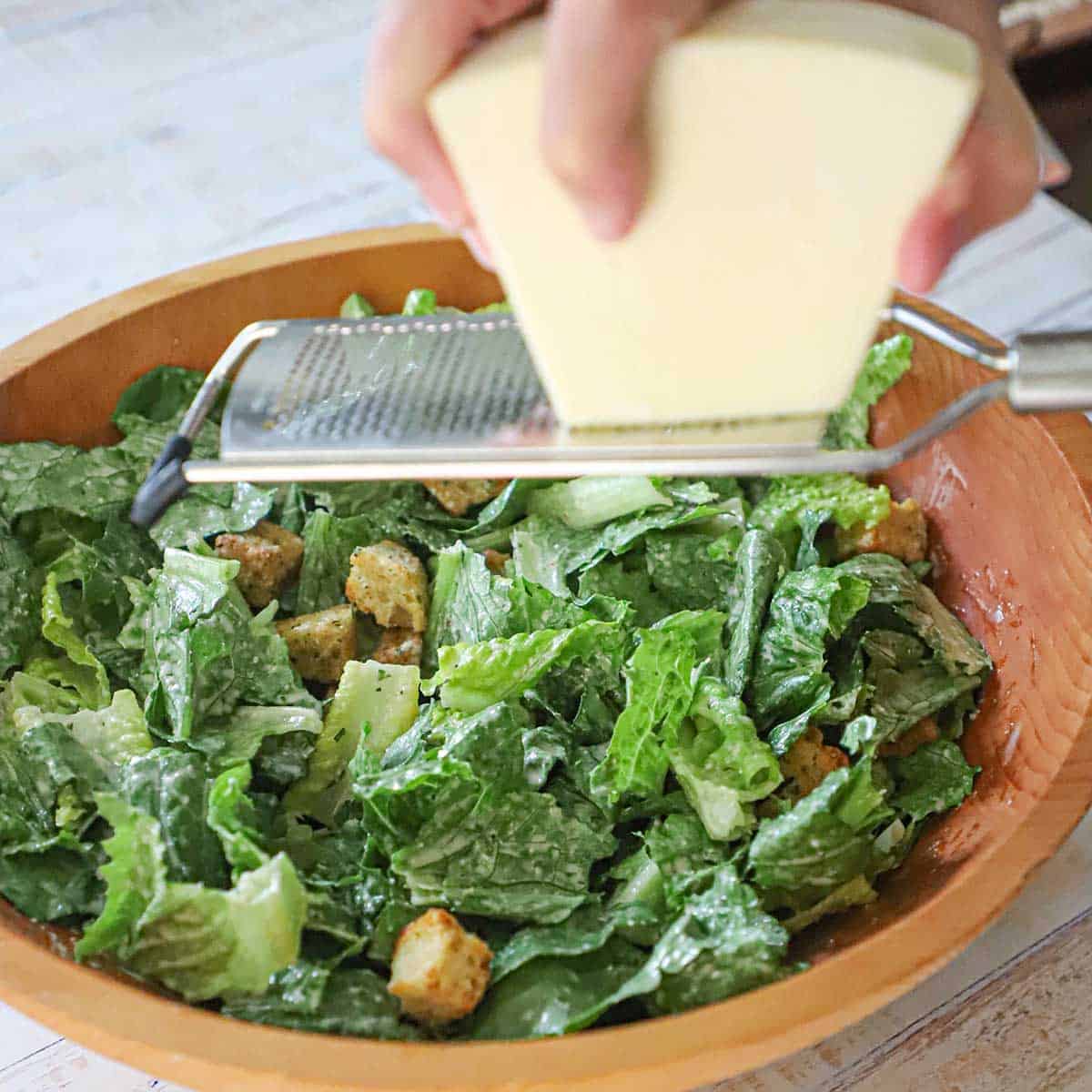A person using a microplane to grate a wedge of Parmesan cheese over the top of a classic Caesar salad that is in a large wooden bowl.