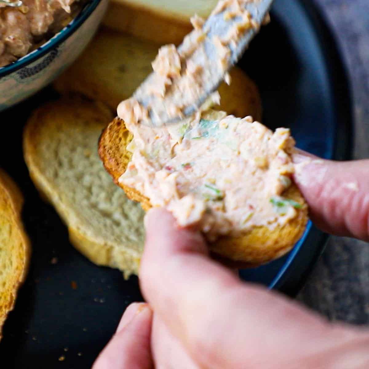 A person holding a toasted slice of a baguette and using a small knife to spread on sun-dried tomato and artichoke dip.