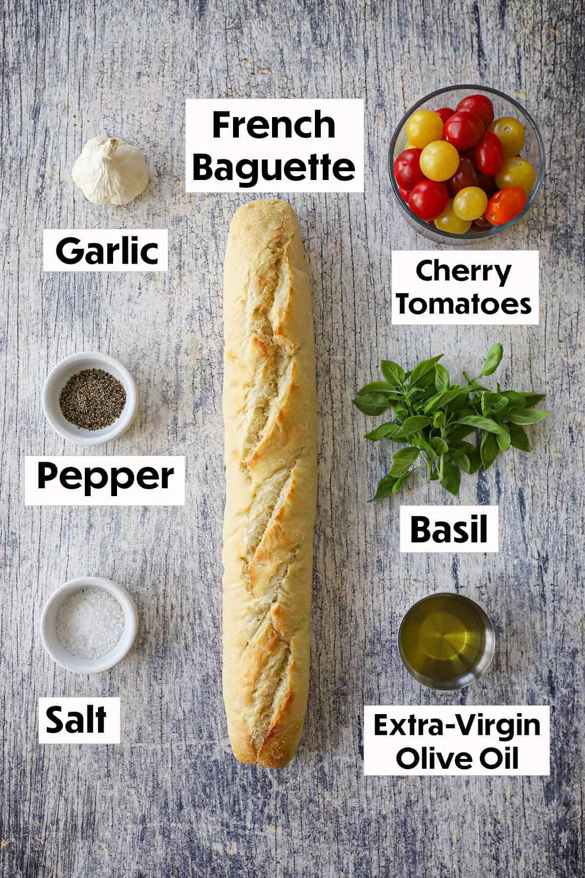 An arrangement of ingredients for Summer bruschetta with tomato and basil on a grey wooden background including a French baguette, garlic, cherry tomatoes, basil, olive oil, salt, and pepper.