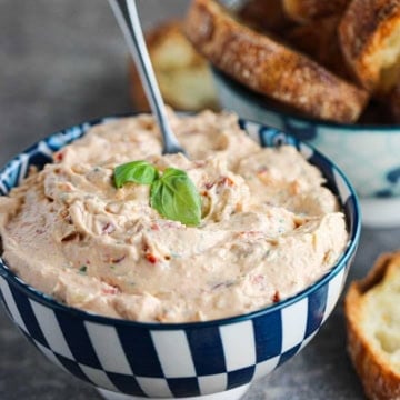 A close-up view of a white and blue checkered bowl filled with sun-dried tomato and artichoke dip with two small basil leaves resting on top.