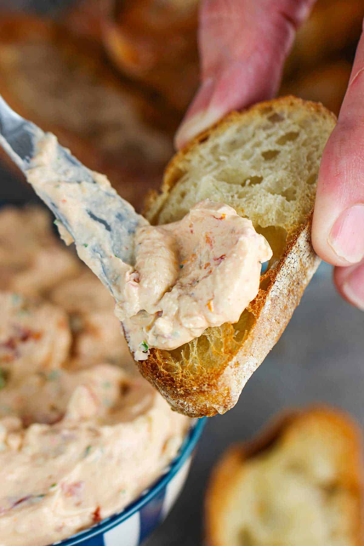 A person using a small butter knife to spread sun-dried tomato and artichoke dip onto a toasted slice of a baguette.
