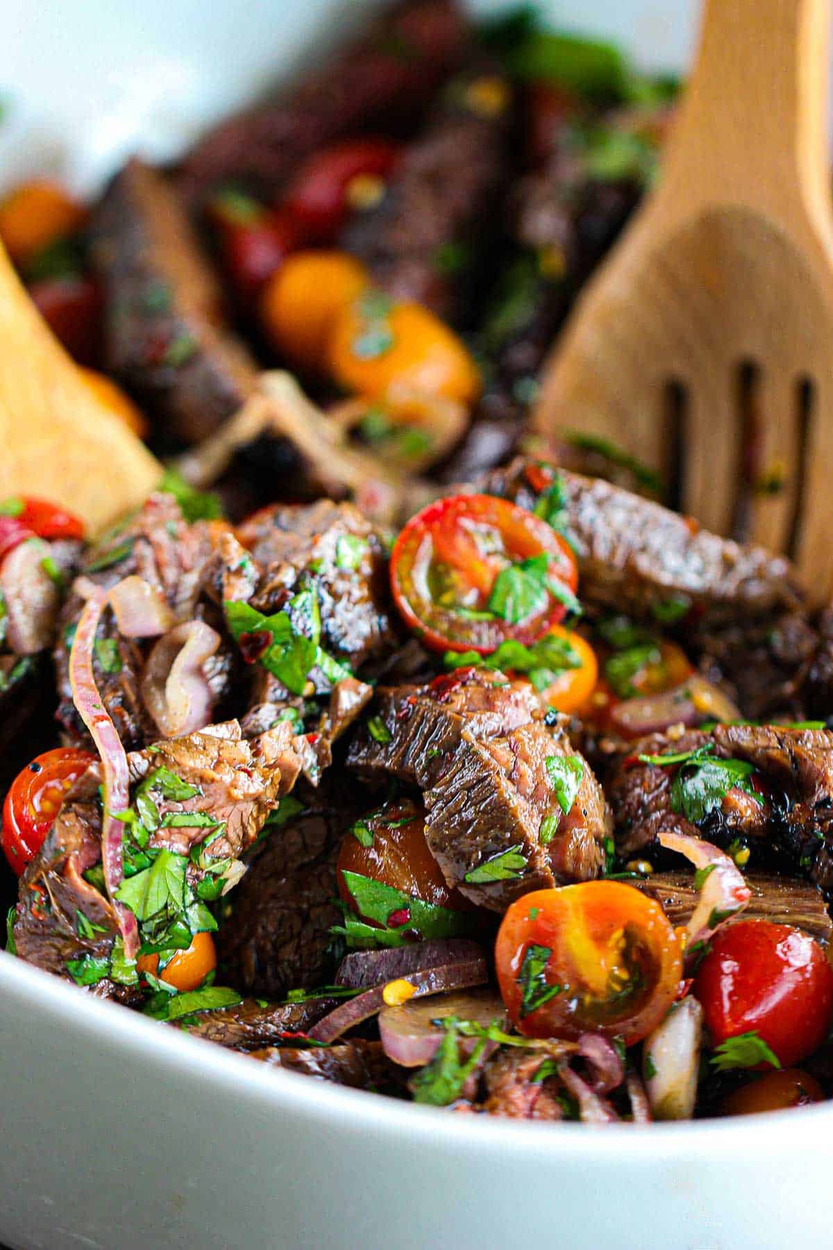 A white bowl filled with Thai beef salad consisting of strips of grilled beef, cherry tomatoes, and herbs all being tossed in a Thai sauce with two wooden spoons.