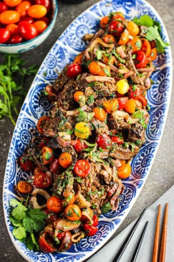 An overhead view of long oval blue and white platter filled with a colorful Thai beef salad with strips of grilled steak, cherry tomatoes, and herbs in a Thai sauce.