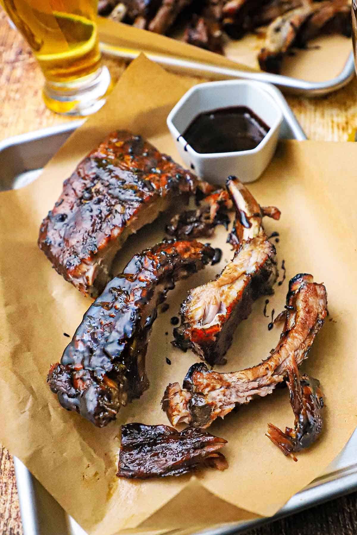 A small baking pan lined with brown paper and topped with balsamic glazed baby back ribs with a small bowl filled with the glaze nearby.