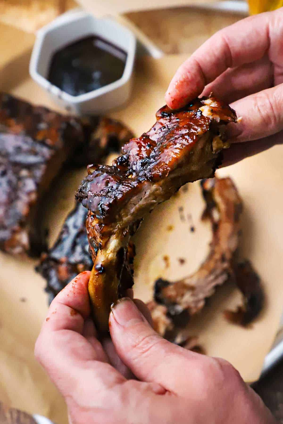 A person holding a balsamic glazed baby back rib over a trey of the entire rack of ribs with a small bowl of the glaze nearby.