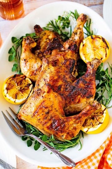 A large oval platter filled with four best grilled chicken quarters with fresh herbs and grilled lemon halves along the perimeter of the dish.