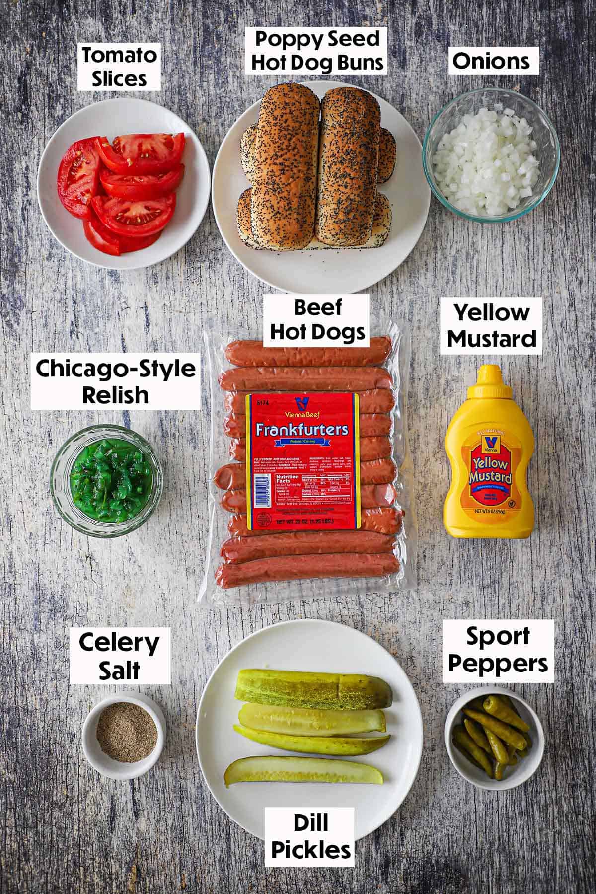 An arrangement of ingredients for Chicago-style hot dogs on a wooden grey background including beef frankfurters, poppyseed buns, tomatoes, onions, green relish, mustard, sport peppers, dill pickle spears, and celery salt.