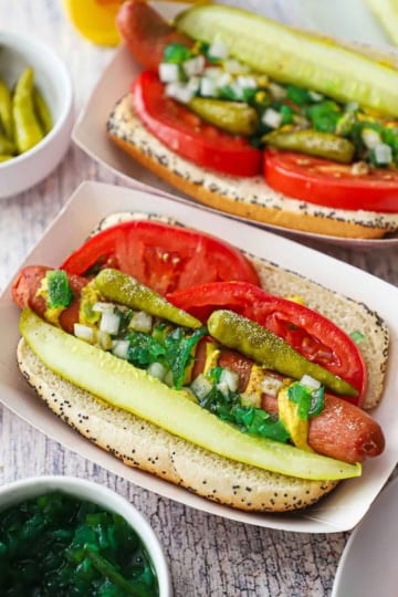 Two Chicago-style hot dogs in paper hot dog holders topped with a spear pickle, relish, peppers, and tomato wedges nestled around the hot dog inside a poppyseed bun.