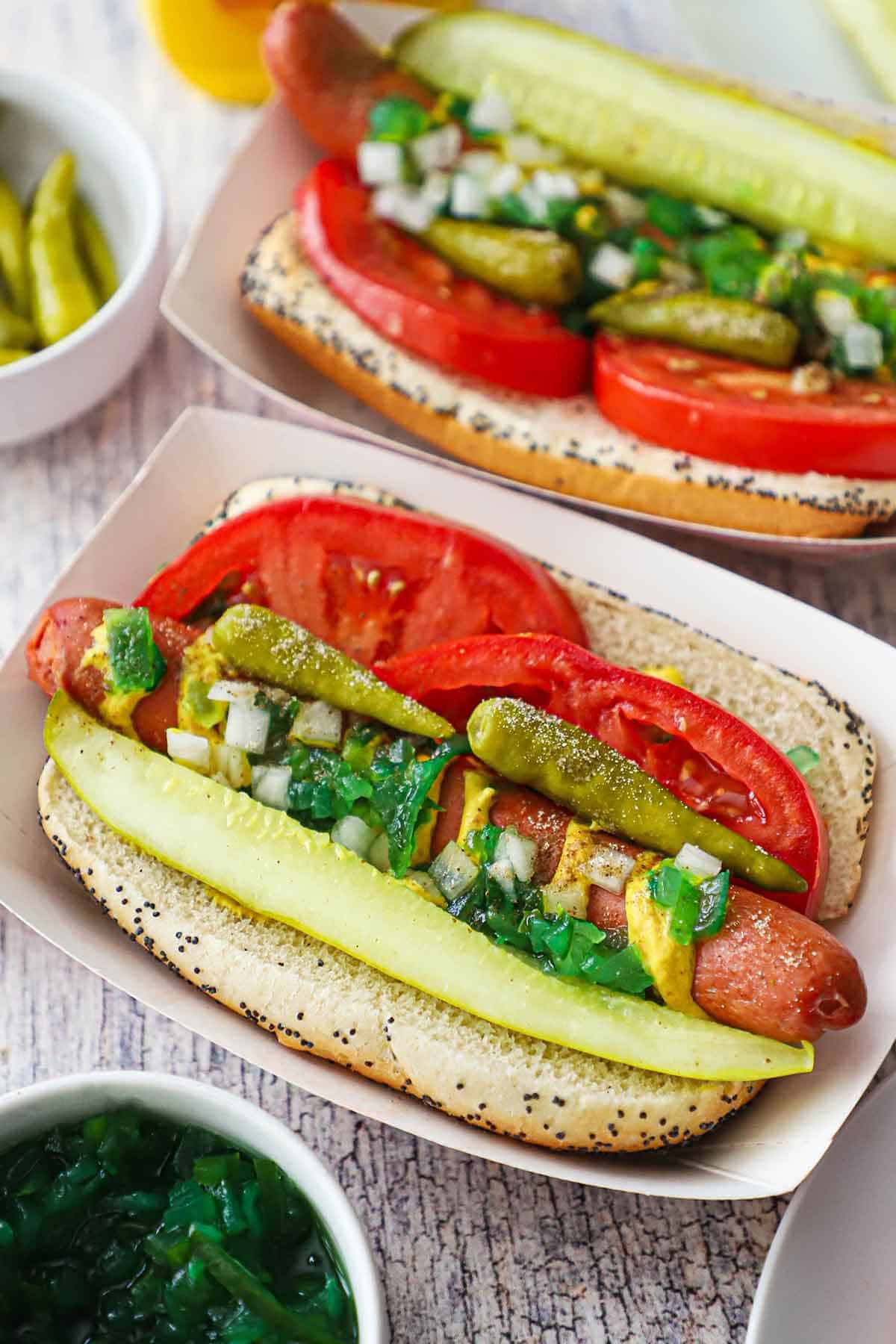 Two Chicago-style hot dogs in paper hot dog holders topped with a spear pickle, relish, peppers, and tomato wedges nestled around the hot dog inside a poppyseed bun.