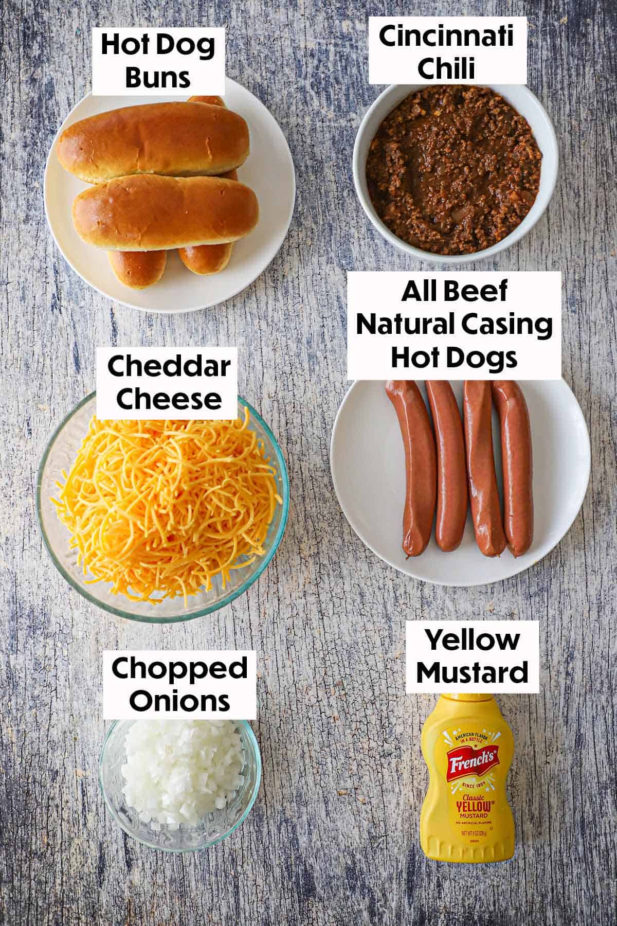 An arrangement of ingredients for Cincinnati Cheese Coney Hot Dogs on a wooden grey background including beef hot dogs, Cincinnati chili, hot dog buns, shredded cheddar cheese, chopped onions, and a bottle of yellow mustard.