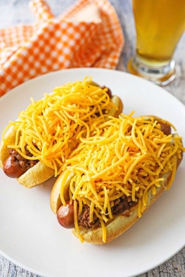 Two Cincinnati Cheese Coney hot dogs sitting on a white dinner plate, both dogs topped with large piles of shredded cheddar cheese.