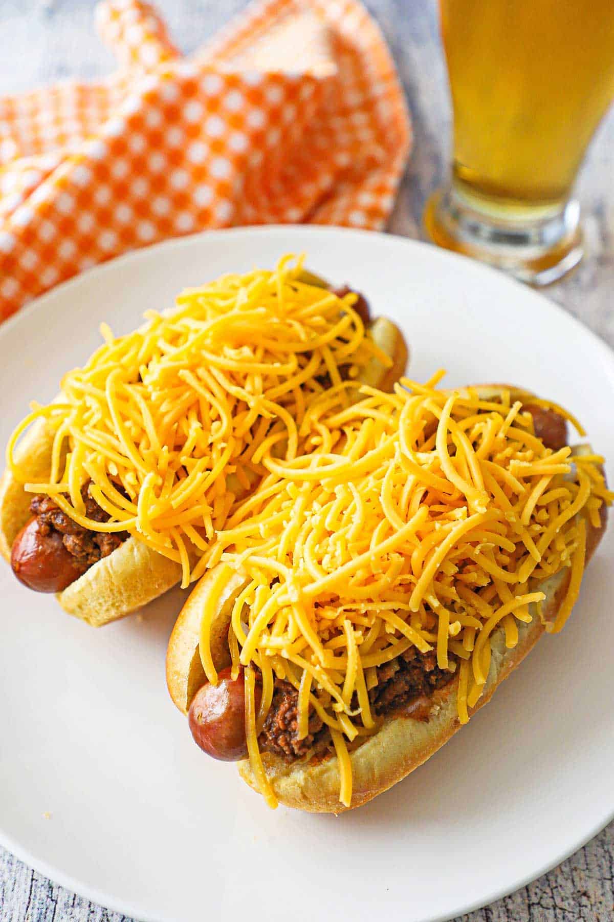 Two Cincinnati Cheese Coney hot dogs sitting on a white dinner plate, both dogs topped with large piles of shredded cheddar cheese.