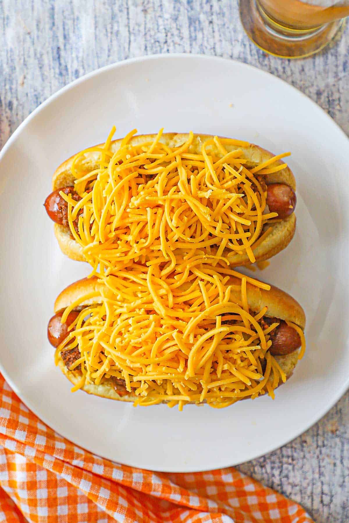 An overhead view of two Cincinnati cheese Coney hot dogs that are both piled high with shredded cheddar cheese on a white plate next to a tall glass of beer.