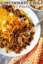 A fork inserted into a large serving of Cincinnati chili on an oval white dinner plate filled with the chill over pasta and topped with a large mound of shredded cheddar cheese and oyster crackers.