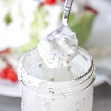 A spoon filled with homemade blue cheese dressing being held over a jar filled with the dressing.