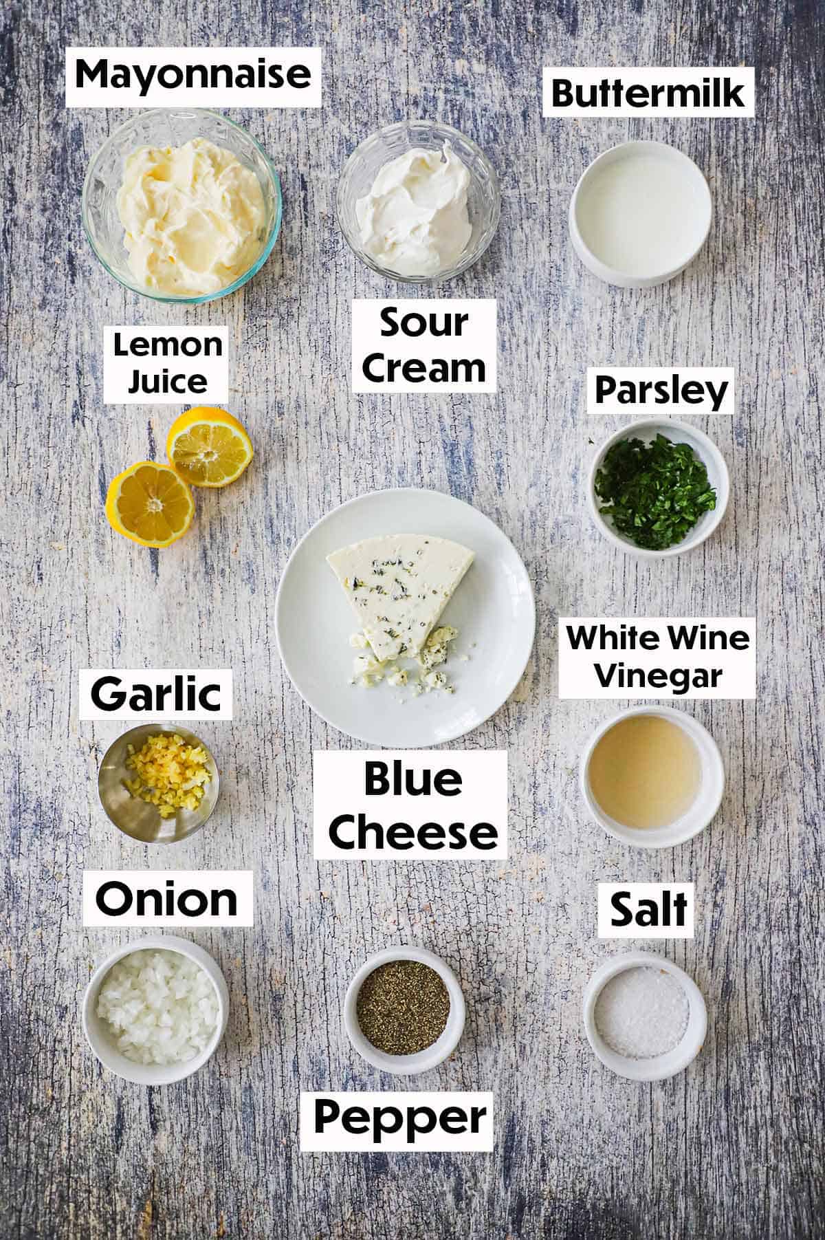 An arrangement of ingredients for homemade blue cheese dressing on a grey wooden background including mayonnaise, sour cream, buttermilk, lemons, blue cheese parsley, garlic, vinegar, onion, salt, and pepper.