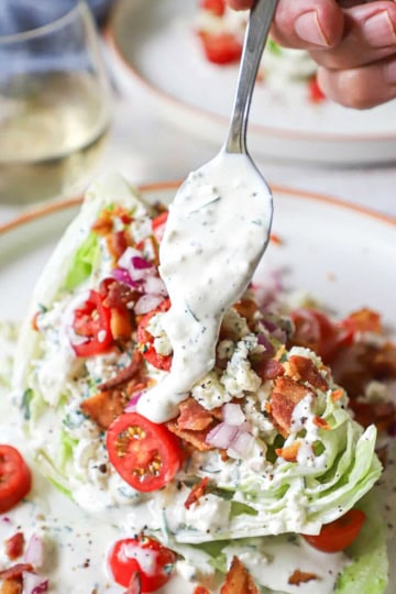 A person transferring homemade blue cheese dressing from a spoon onto a wedge salad that includes iceberg lettuce, chopped tomatoes, crumbled bacon, and chopped onion.