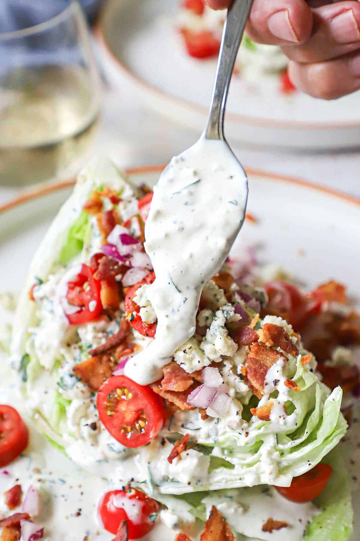 A person transferring homemade blue cheese dressing from a spoon onto a wedge salad that includes iceberg lettuce, chopped tomatoes, crumbled bacon, and chopped onion.