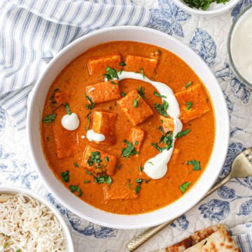 A white serving bowl filled with paneer butter masala and topped with streaks of cream and garnished with chopped cilantro.