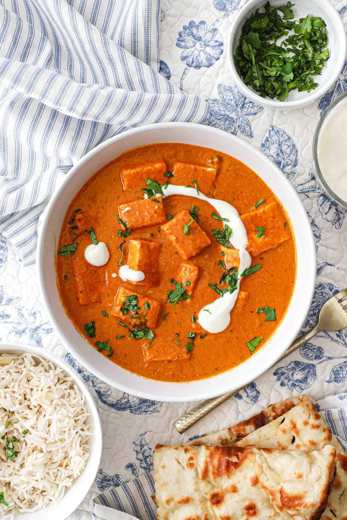 A white serving bowl filled with a serving of paneer butter masala with a bowl of basmati rice, a stack of naan, and a small bowl of chopped cilantro nearby.