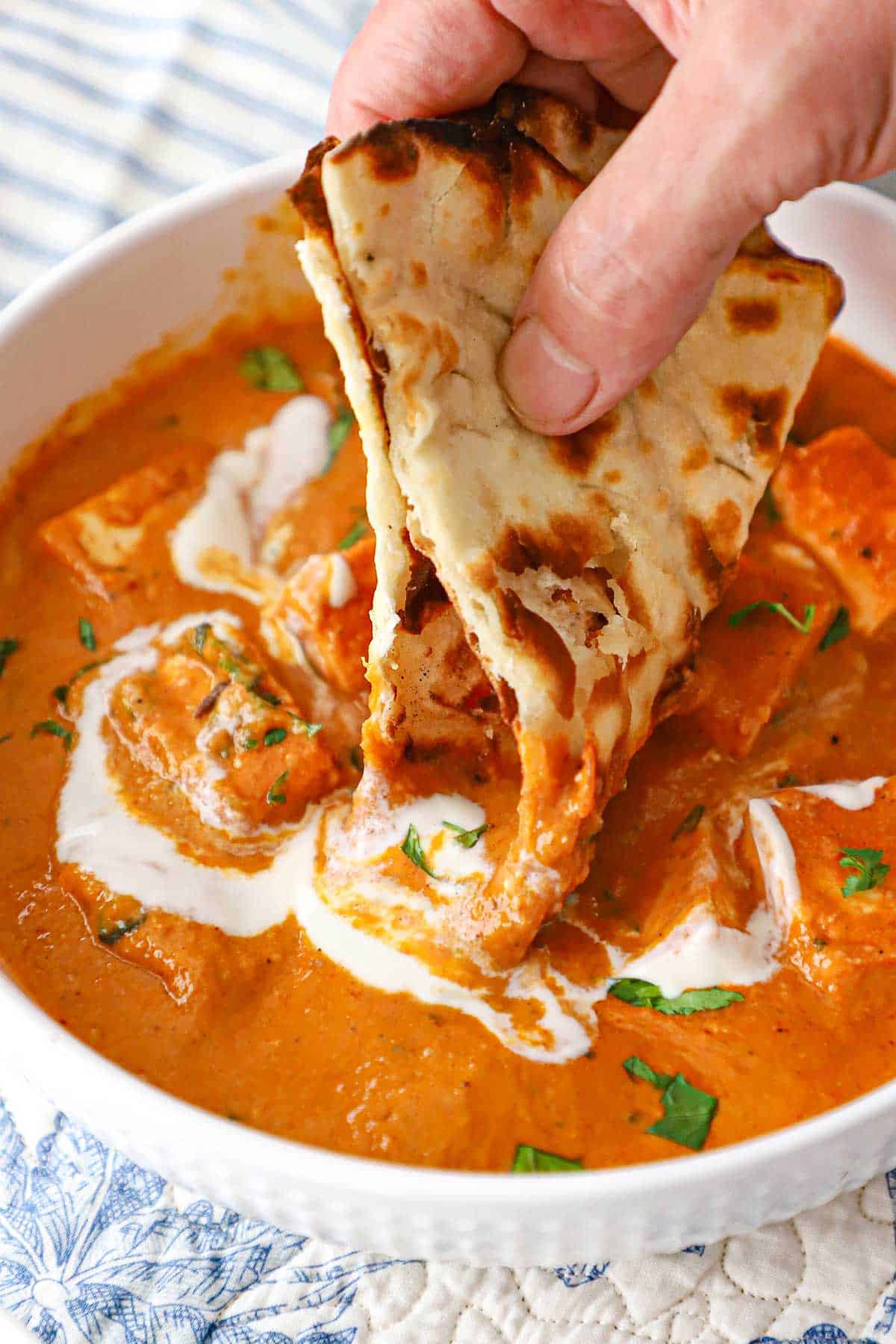 A person plunging a folded piece of naan into a white bowl filled with a serving of panner butter masala.
