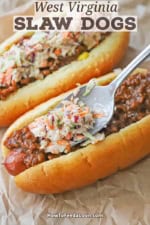 A person using a spoon to add a layer of coleslaw over a layer of hot dog sauce that is resting on a hot dog in a hot dog bun on crumpled brown wax paper.