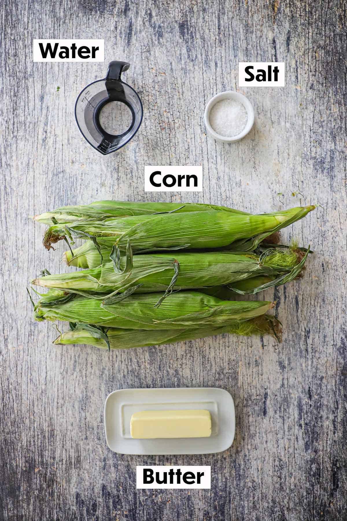 An arrangement of ingredients for slow cooker corn on the cob on a grey wooden background including ears of corn in their husks, a stick of butter, salt, and water.