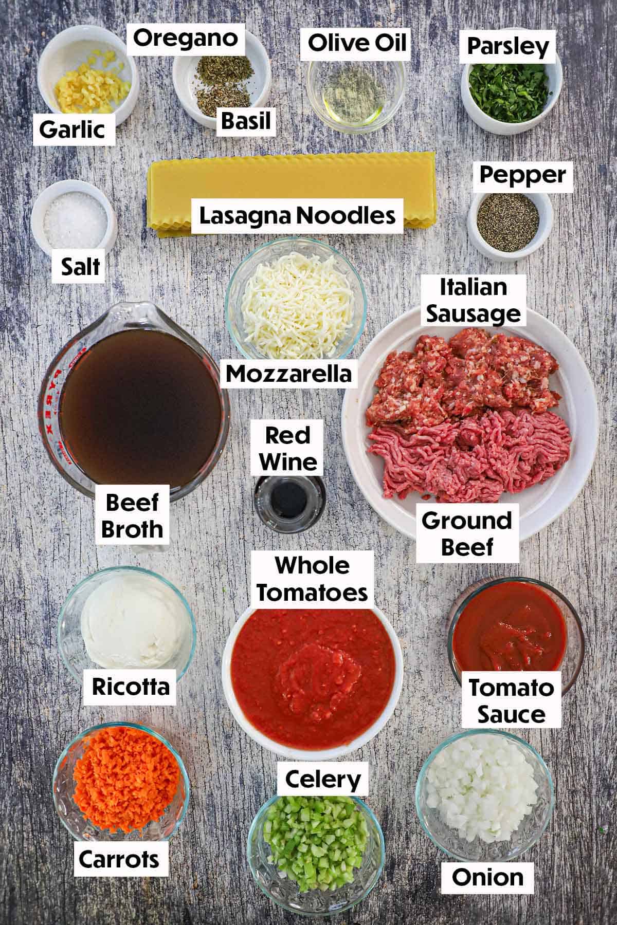 An arrangement of ingredients for slow cooker lasagna soup on a wooden grey background including ground beef, Italian sausage garlic, beef broth, tomatoes, ricotta cheese, vegetables, and herbs.