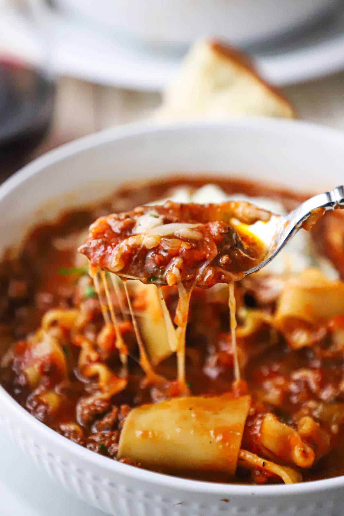 A spoon being used to raise a spoonful of lasagna soup from a white bowl with stretches of cheese visible between spoon and the soup.