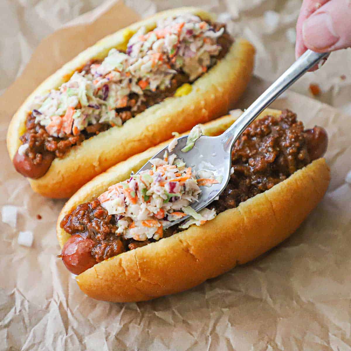 A person using a spoon to add a layer of coleslaw over a layer of hot dog sauce that is resting on a hot dog in a hot dog bun on crumpled brown wax paper.