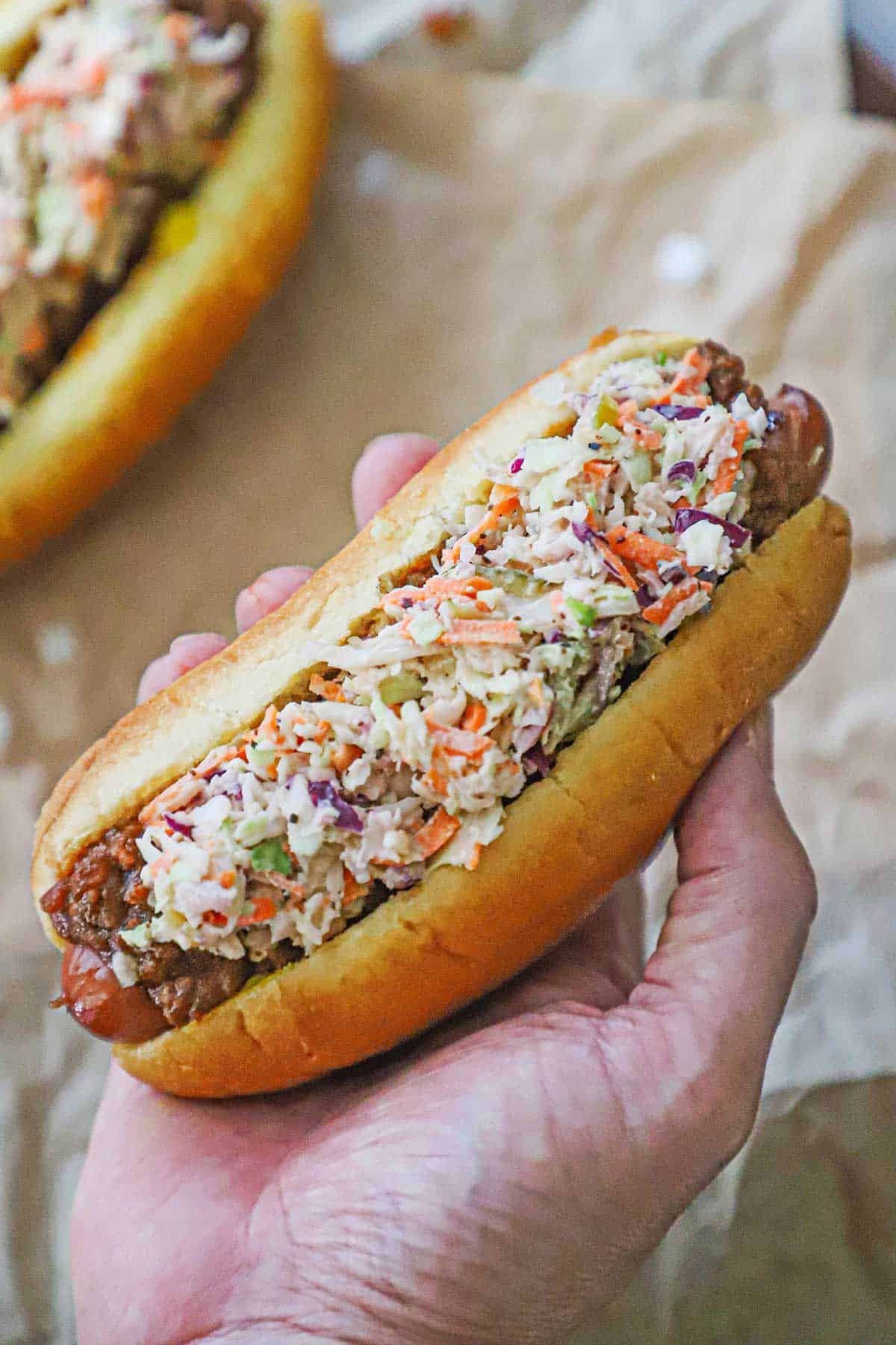 A person holding a West Virginia Slaw Dog in the palm of his hand over another slaw dog that is resting on a crumpled piece of brown paper.