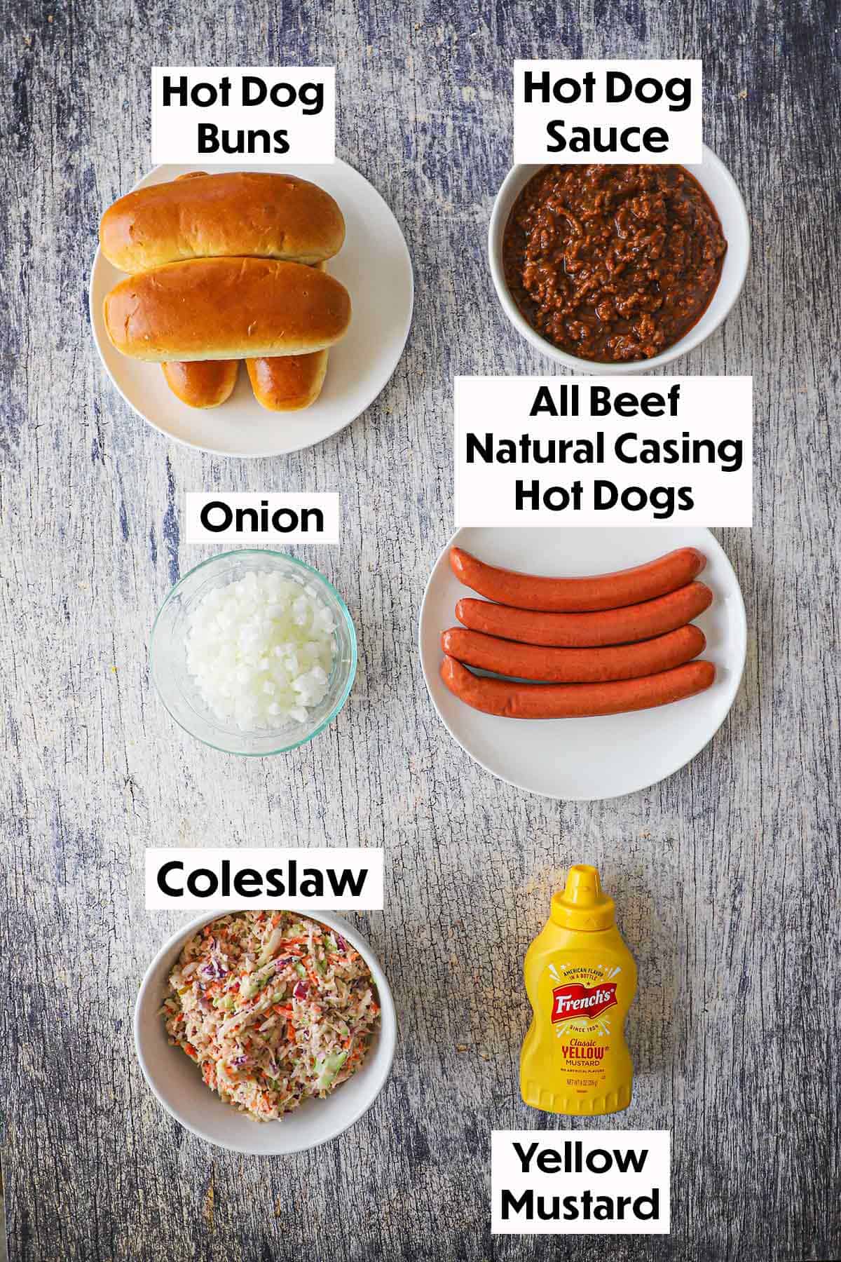 An arrangement of ingredients for West Virginia Slaw Dogs on a wooden grey background including all beef natural casing hot dogs, hot dog sauce, hot dog buns, chopped onion, coleslaw, and a bottle of yellow mustard.