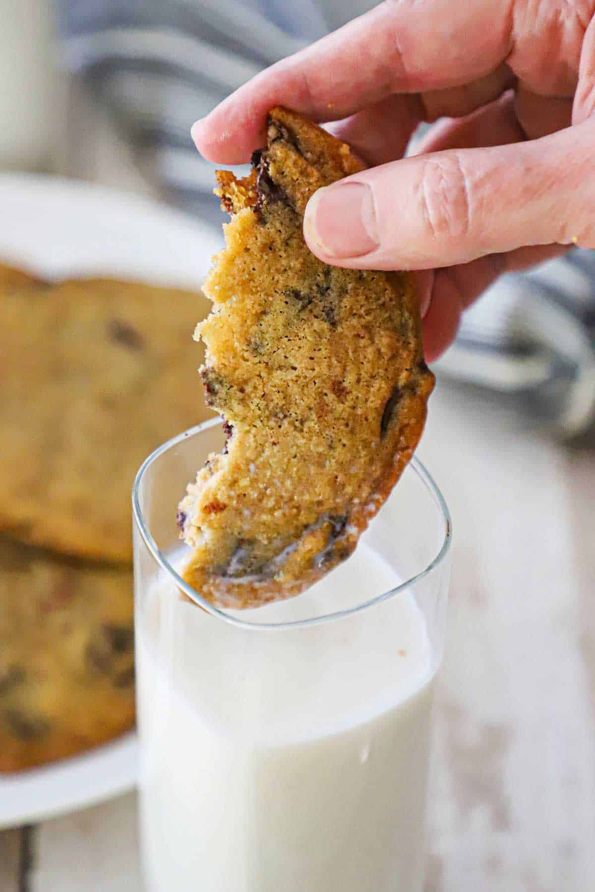 A person dunking half of a bakery-style chocolate chip cookie into a tall glass of milk next to a plate of the cookies.