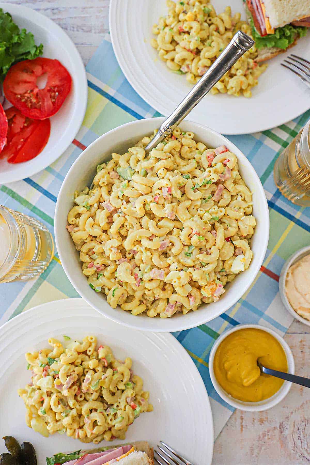 An overhead view of a white serving bowl filled with classic macaroni salad with two plates nearby containing more macaroni salad and a ham sandwich.